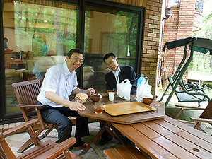 「峠の釜めし」を食べて幸せそうな二人とかたや左後方では必死にグランドピアノを納品している青い制服の伸和ピアノさん